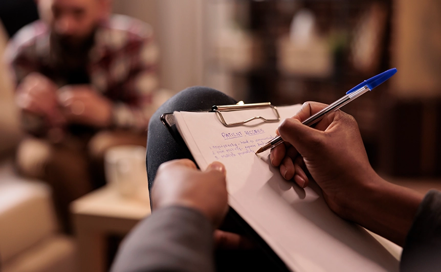 Person Taking Notes During Counseling Session with Client in Background