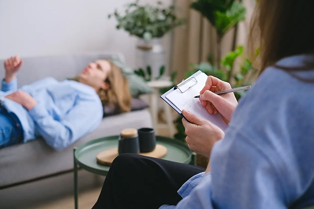 Person writing on clipboard beside individual lying on couch in therapy session.